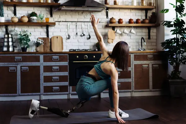 Female with leg prosthesis balances in yoga position on mat at home. Lady performs exercises on floor of kitchen demonstrating balance and strength