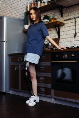 Yapay bacaklı esmer kadın kollarını kavuşturarak mutfakta duruyor. Özürlü bir kadın, rahat bir mutfakta kameraya poz vererek kendine güveniyor.
