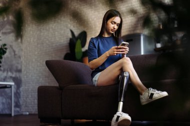 Woman with disability sits on sofa and types messages on smartphone. Attractive lady with prosthetic leg engaged in lively chat with boyfriend on mobile phone clipart