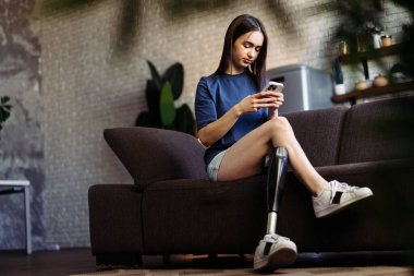 Engelli bir kadın kanepeye oturur ve akıllı telefondan mesajlar yazar. Protez bacaklı çekici bayan cep telefonuyla erkek arkadaşıyla canlı sohbete daldı.