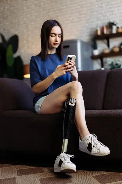 Woman with disability sits on sofa and types messages on smartphone. Attractive lady with prosthetic leg engaged in lively chat with boyfriend on mobile phone