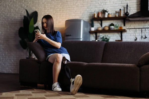 Young woman with disability shares stories with friend using phone. Lady with prosthetic leg in casual attire sits on soft couch and types messages on smartphone