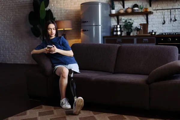 Young woman with disability shares stories with friend using phone. Lady with prosthetic leg in casual attire sits on soft couch and types messages on smartphone
