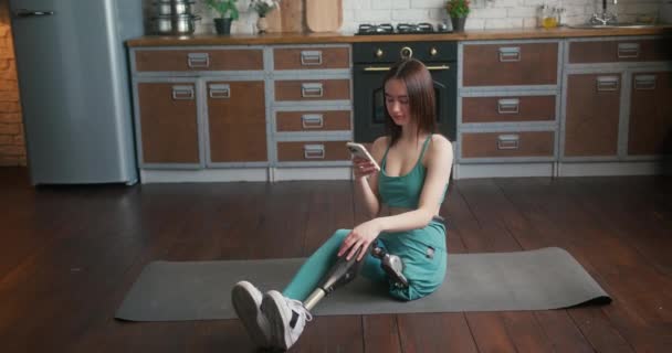 Kobieta Protezą Nóg Siedzi Macie Fitness Wiadomości Przez Telefon Młoda — Wideo stockowe