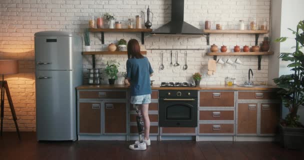 義足を持つ女性は 食糧を準備するためにガス調理器に鍋を置きます 自宅で近代的なキッチンで新しいレシピで障害実験をしている若い女性 — ストック動画