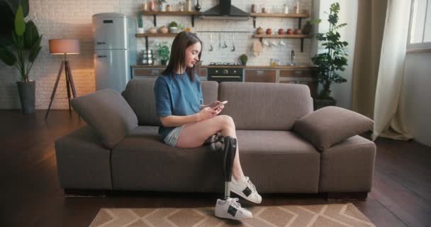Kobieta Niepełnosprawna Siedzi Kanapie Pisze Wiadomości Smartfonie Atrakcyjna Kobieta Protezą — Wideo stockowe