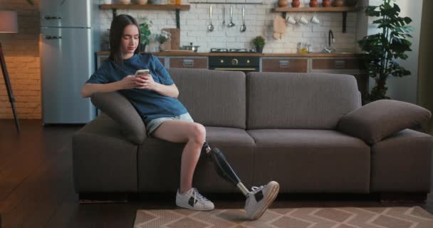 障害を持つ若い女性は 電話を使って友人と物語を共有しています カジュアルな服装で義足を持つ女性はソフトソファに座り スマートフォンでメッセージをタイプします — ストック動画