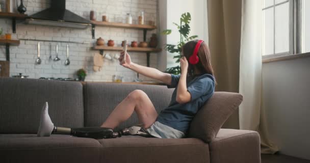 障害のある女性は 柔らかいソファーに座っているヘッドフォンを介して音楽を好むようにリストします 義足の女性は瞬間をキャプチャするためにセルフィーを取る — ストック動画