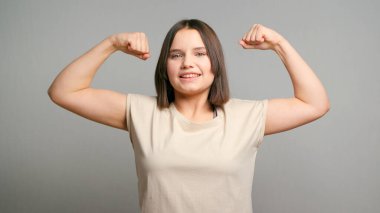 Genç güçlü Latin kız kendini mutlu, tatmin olmuş ve güçlü hissediyor, kaslı ve formda, gri arka planda güçlü görünüyor..