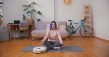 Elektrikli süpürge robotu etrafta dolaşırken meditasyon yapmaya çalışan Latin bir genç kız. Kapalı alandaki bitkilerle iç içe. Çevrimiçi fitness, kentsel orman konsepti.