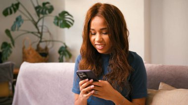 Latin bayan evdeki cep telefonundan mesajlar yazıyor. Kanepede oturan, akıllı telefondan gülümseyen İspanyol kadın mesajları. Karışık ırk toleransı