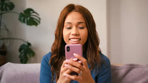 拉丁女人在家里用手机发短信笑 坐在沙发上的智能手机上快乐的拉美裔女性 混合种族容忍 图库照片