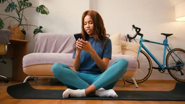 西班牙裔女士坐在瑜伽垫上 用手机发短信 年轻的拉丁女士在家庭训练中使用社交媒体休息 混合种族容忍 图库图片