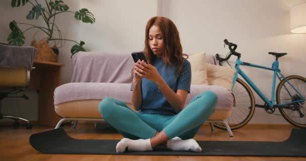 西班牙裔女士坐在瑜伽垫上 用手机发短信 年轻的拉丁女士在家庭训练中使用社交媒体休息 混合种族容忍 — 图库视频影像