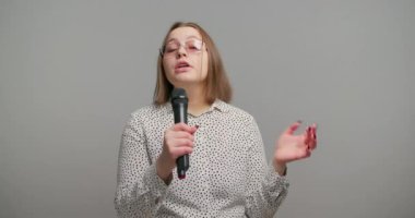 Gülümseyen kadın gri arka planda yavaşça mikrofonla konuşuyor. Kız, seyirciyi cezbedici varlığıyla kolayca kendine çekiyor.