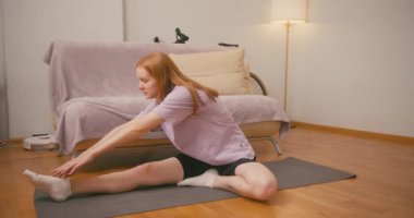 Kızıl saçlı kadın evde yerde bacak esneme hareketleri yapıyor. Sporcu kadın, ev egzersizi sırasında esneklik ve kas tonunu geliştirir