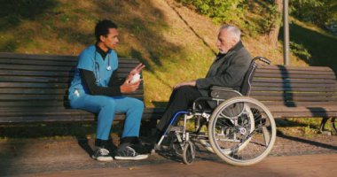 İspanyol doktor tekerlekli sandalyedeki engelli arkadaşla ilgileniyor. Latin sıhhiyeci, yaşlı adama sonbahar parkında rehabilitasyonda yardım ediyor. Sağlık, sakatlık, hareketlilik, arkadaşlık.