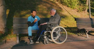 İspanyol doktor tekerlekli sandalyedeki engelli arkadaşla ilgileniyor. Latin sıhhiyeci, yaşlı adama sonbahar parkında rehabilitasyonda yardım ediyor. Sağlık, sakatlık, hareketlilik, arkadaşlık.