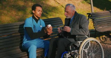İspanyol doktor tekerlekli sandalyedeki engelli arkadaşla ilgileniyor. Latin sıhhiyeci, yaşlı adama sonbahar parkında rehabilitasyonda yardım ediyor. Sağlık, sakatlık, hareketlilik, arkadaşlık.
