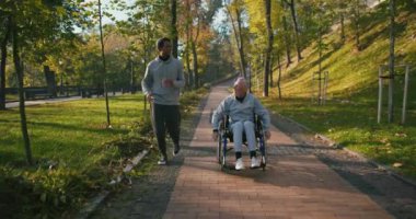 İspanyol adam tekerlekli sandalyedeki engelli arkadaşla ilgileniyor. Sonbahar parkında birlikte eğlenen arkadaşlar. Sağlık, sakatlık, hareketlilik, arkadaşlık.