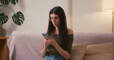 Oturma odasındaki koltukta oturan genç bir kadın cep telefonuna mesaj yazıyor. Neşeli İspanyol bayan internetten alışveriş yapıyor. Evde sipariş bekliyor.