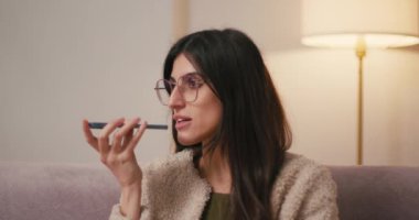 İspanyol kadın evdeki koltukta oturan sesli mesajları kaydediyor. Sarı saçlı, yün ceketli esmer kadın akıllı telefon kullanarak heyecan verici bir hikayeyi paylaşıyor.