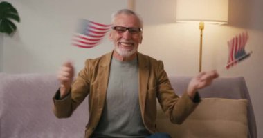 Mutlu kıdemli adam aktif olarak evdeki koltukta oturan ABD bayraklarını sallıyor. Gözlüklü ve ceketli gri saçlı Arap emeklisi ulusal bayramı kutluyor