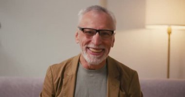 Mutlu kıdemli adam aktif olarak evdeki koltukta oturan ABD bayraklarını sallıyor. Gözlüklü ve ceketli gri saçlı Arap emeklisi ulusal bayramı kutluyor