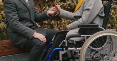 Koca, tekerlekli sandalyede oturan emekli bir eşin ellerini sıvazlıyor. Yaşlı evli çift, sonbahar parkında boş vakit geçirmeyi sevdiklerini gösteriyor