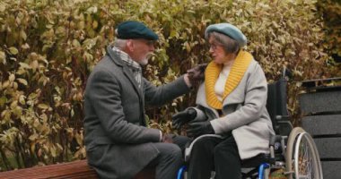 Yaşlı koca bankta dinleniyor ve karısı tekerlekli sandalyede oturuyor. Paltolu ve şapkalı mutlu emekli çift doğada eğlenerek vakit geçiriyor.