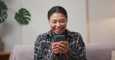 Gülen Latin Amerikalı kadın oturma odasında akıllı telefondan mesaj gönderiyor. Esmer kadın sosyal medyada internette sohbet ediyor evde komik mimler yiyor.