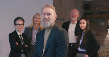 Gülümseyen sakallı kıdemli adam kollarını kavuşturmuş dururken, çok ırklı iş adamları ofiste ayakta duruyor. Çeşitli işçiler toplantı odasında işbirliği yapıyorlar.