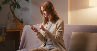 Kızıl saçlı kadın, apartmandaki koltukta oturan akıllı telefondan sosyal medyayı kaydırıyor. Genç bayan, akşamları işten sonra dinlenirken dijital içeriğe göz atıyor