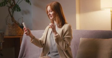 Gülümseyen kızıl saçlı kadın evdeki kanepede oturan akıllı telefondan video konuşmalarıyla konuşuyor. Neşeli kadın erkek arkadaşıyla sanal iletişim kuruyor.