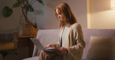 Oturma odasındaki kanepede oturan dizüstü bilgisayarda odaklanmış iş kadını tipleri. Kızıl saçlı, uzun boylu genç bir kadın akşamları evden çalışırken günlüğüne not alıyor.