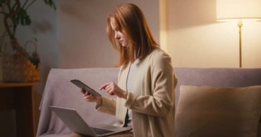 Kızıl saçlı kadın, oturma odasındaki kanepede oturan tablet ve dizüstü bilgisayarla çalışıyor. Gevşek saçlı genç bir kadın iş akışını optimize etmek için modern aletler kullanıyor.