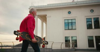 Aktif, mutlu, yaşlı, kırmızı tişörtlü kaykaycı gün batımından sonra şehir merkezinde kaykayla geziyor. Yaşlılar özgürlük ruhu kavramı. Yavaş çekim.