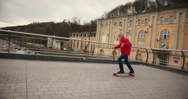 Aktif, mutlu, yaşlı kaykaycı kırmızı tişörtlü şehir merkezinde gün batımında kaykaya biniyor. Yaşlılar özgürlük ruhu kavramı. Yavaş çekim.