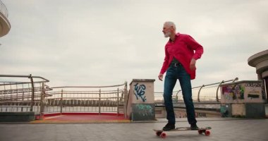 Aktif, mutlu, yaşlı kaykaycı kırmızı tişörtlü şehir merkezinde gün batımında kaykaya biniyor. Yaşlılar özgürlük ruhu kavramı. Yavaş çekim.