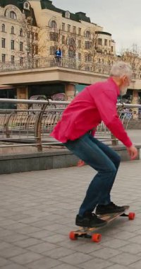 Aktif, mutlu, yaşlı kaykaycı kırmızı tişörtlü şehir merkezinde gün batımında kaykaya biniyor. Yaşlılar özgürlük ruhu kavramı. Yavaş çekim.