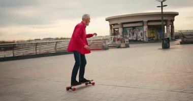 Aktif, mutlu, yaşlı kaykaycı kırmızı tişörtlü şehir merkezinde gün batımında kaykaya biniyor. Yaşlılar özgürlük ruhu kavramı. Yavaş çekim.