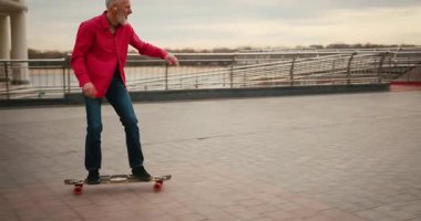 Aktif, mutlu, yaşlı kaykaycı kırmızı tişörtlü şehir merkezinde gün batımında kaykaya biniyor. Yaşlılar özgürlük ruhu kavramı. Yavaş çekim.