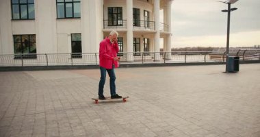 Aktif, mutlu, yaşlı kaykaycı kırmızı tişörtlü şehir merkezinde gün batımında kaykaya biniyor. Yaşlılar özgürlük ruhu kavramı. Yavaş çekim.