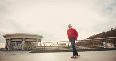 Aktif, mutlu, yaşlı kaykaycı kırmızı tişörtlü şehir merkezinde gün batımında kaykaya biniyor. Yaşlılar özgürlük ruhu kavramı. Yavaş çekim.