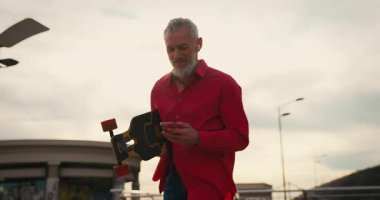 Aktif, mutlu, yaşlı, kırmızı tişörtlü kaykaycı gün batımından sonra şehir merkezinde kaykayla geziyor. Yaşlılar özgürlük ruhu kavramı. Yavaş çekim.