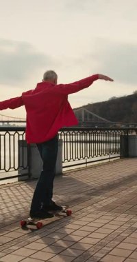 Aktif, mutlu, yaşlı kaykaycı kırmızı tişörtlü şehir merkezinde gün batımında kaykaya biniyor. Yaşlılar özgürlük ruhu kavramı. Yavaş çekim.