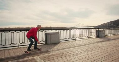 Aktif, mutlu, yaşlı kaykaycı kırmızı tişörtlü şehir merkezinde gün batımında kaykaya biniyor. Yaşlılar özgürlük ruhu kavramı. Yavaş çekim.