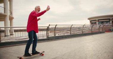 Aktif, mutlu, yaşlı kaykaycı kırmızı tişörtlü şehir merkezinde gün batımında kaykaya biniyor. Yaşlılar özgürlük ruhu kavramı. Yavaş çekim.