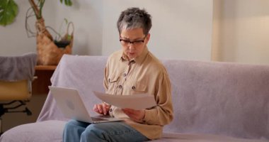 Latin kadın evdeki kanepede dizüstü bilgisayarla otururken istatistiksel verileri gözden geçiriyor. Kıdemli bayan, iş belgelerini konsantre bir ifadeyle analiz eder.