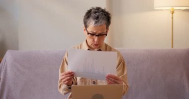 Latin kadın evdeki kanepede dizüstü bilgisayarla otururken istatistiksel verileri gözden geçiriyor. Kıdemli bayan, iş belgelerini konsantre bir ifadeyle analiz eder.